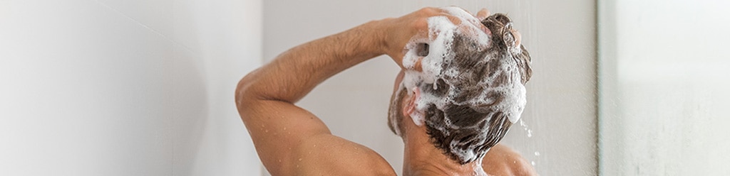 Man wast zijn haren met schuimende shampoo onder de douche